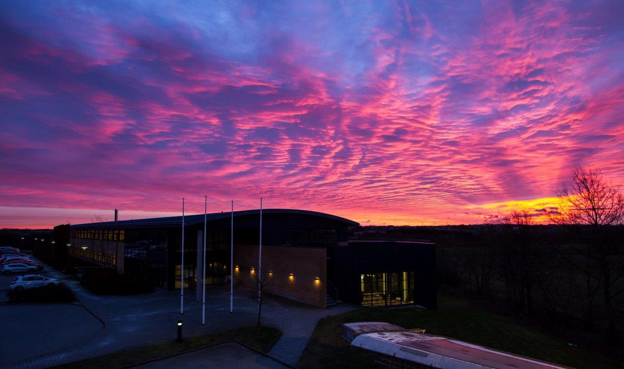Vibrant sunset with pink and purple hues over a building, featuring several flagpoles in the foreground.