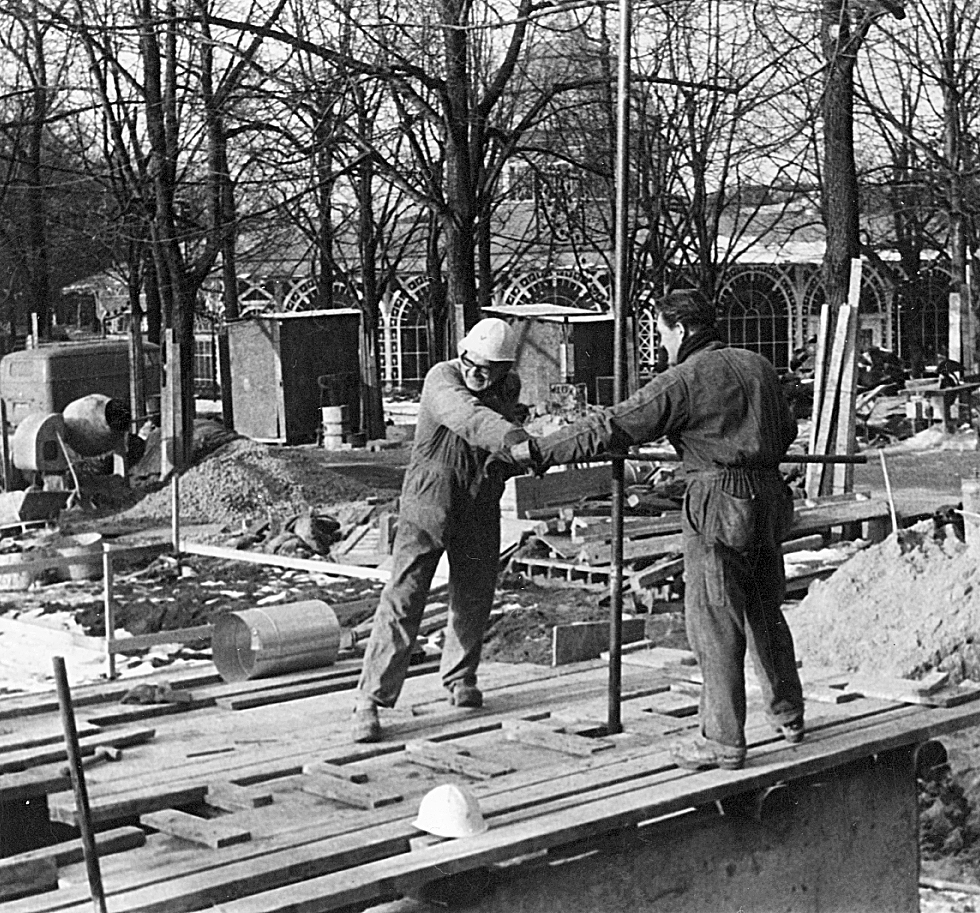 Two construction workers in winter gear collaborate on a building site, handling materials and equipment amidst a snowy landscape.