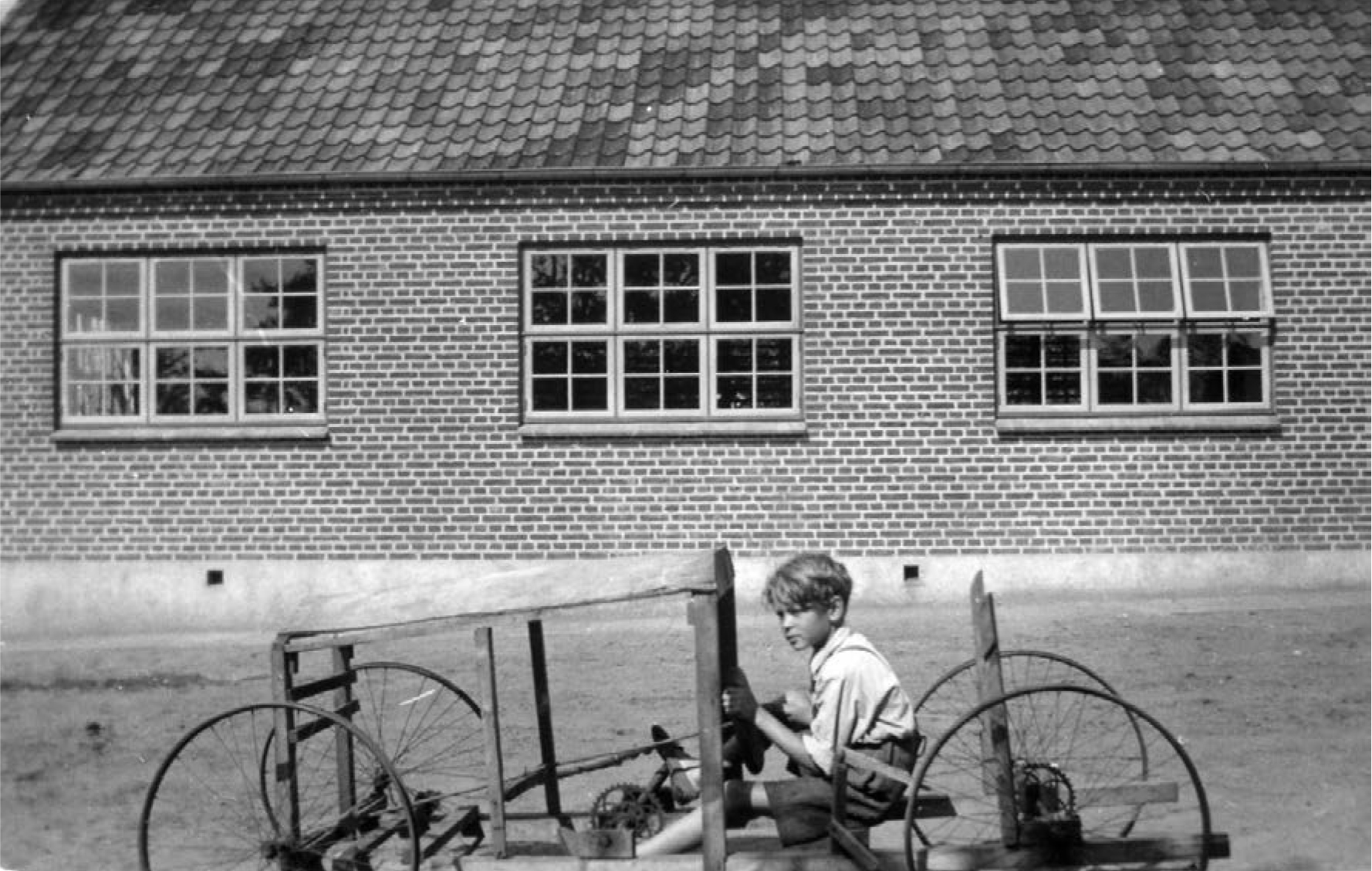 A boy sits on a wooden cart with wheels, focused on a task, in front of a brick building with multiple windows.