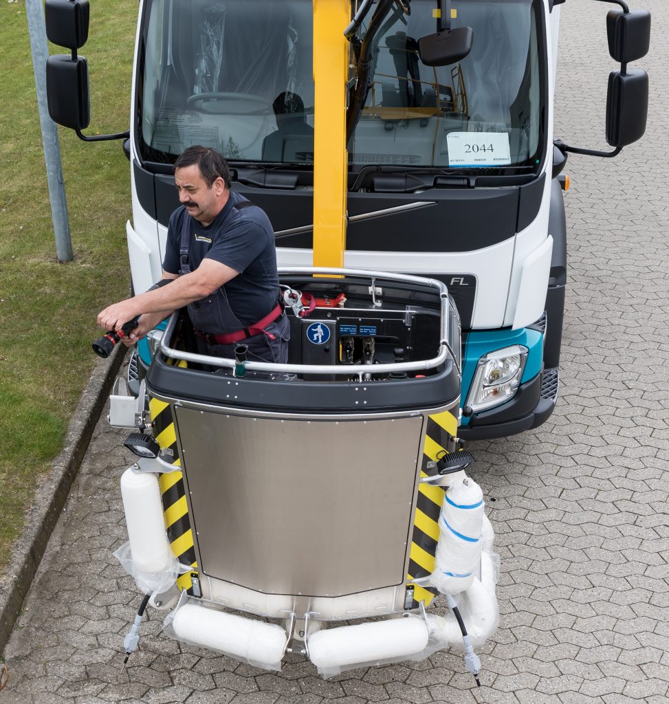 A worker operates a lifting device attached to a truck, featuring safety harnesses and a control panel.