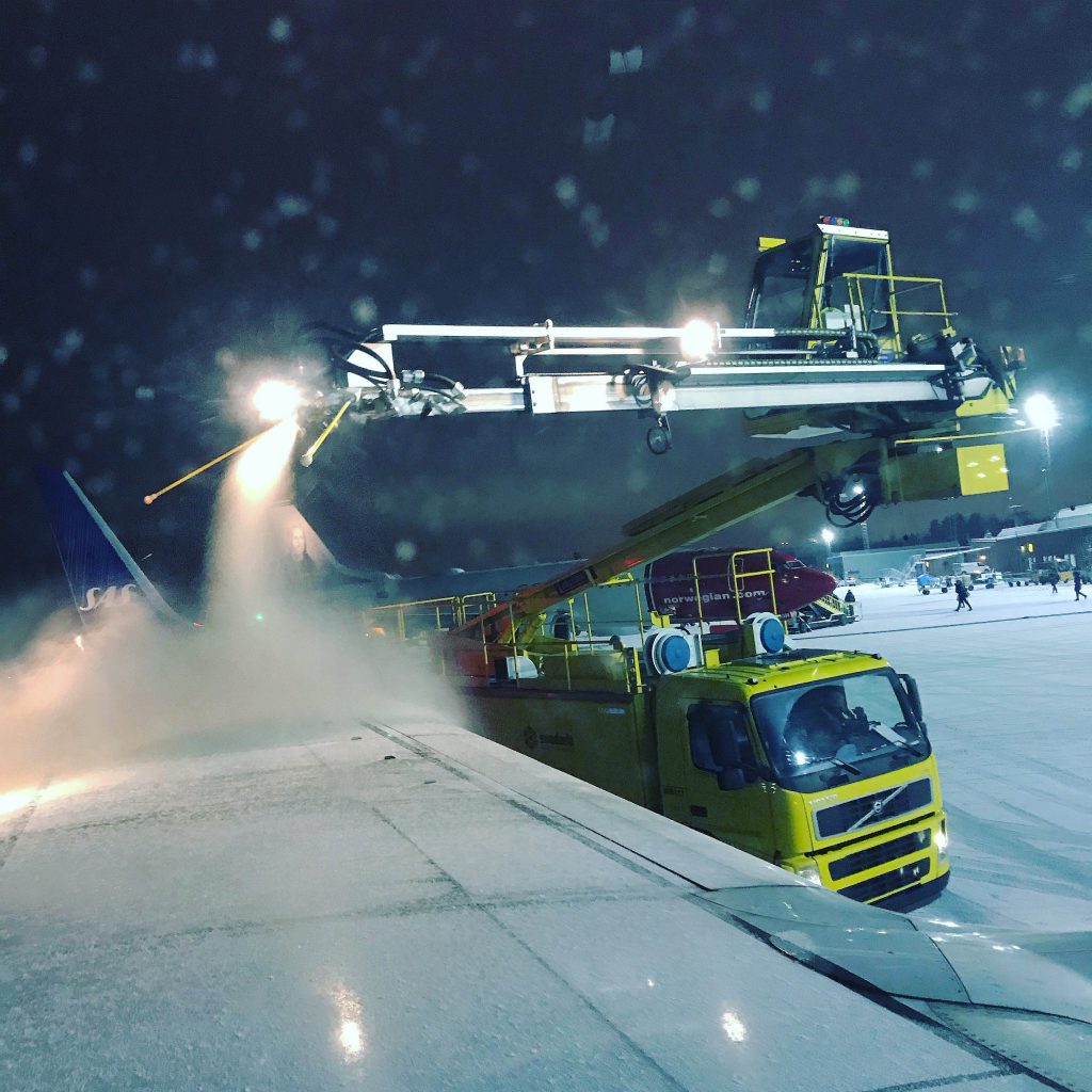 Snow removal vehicle spraying de-icing fluid on an airplane wing during nighttime operations in snowy conditions.