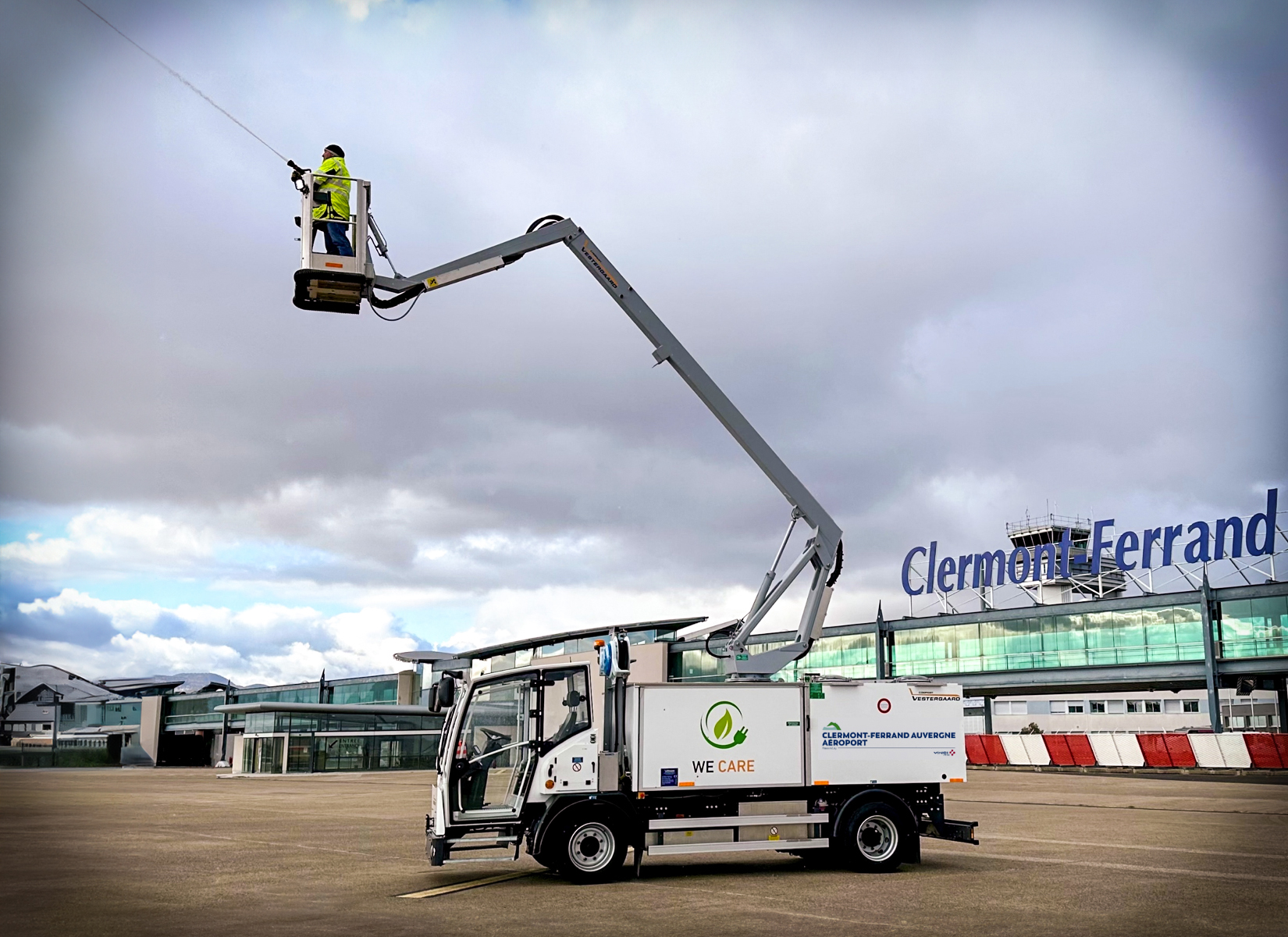 A worker in a lift bucket is inspecting overhead lines near Clermont-Ferrand airport, with a cloudy sky above.
