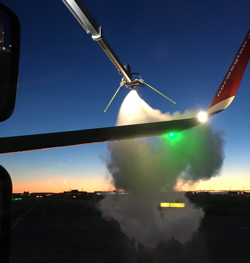 Aircraft de-icing process at dawn, with a spray nozzle releasing fluid onto the wing, creating a cloud of mist.