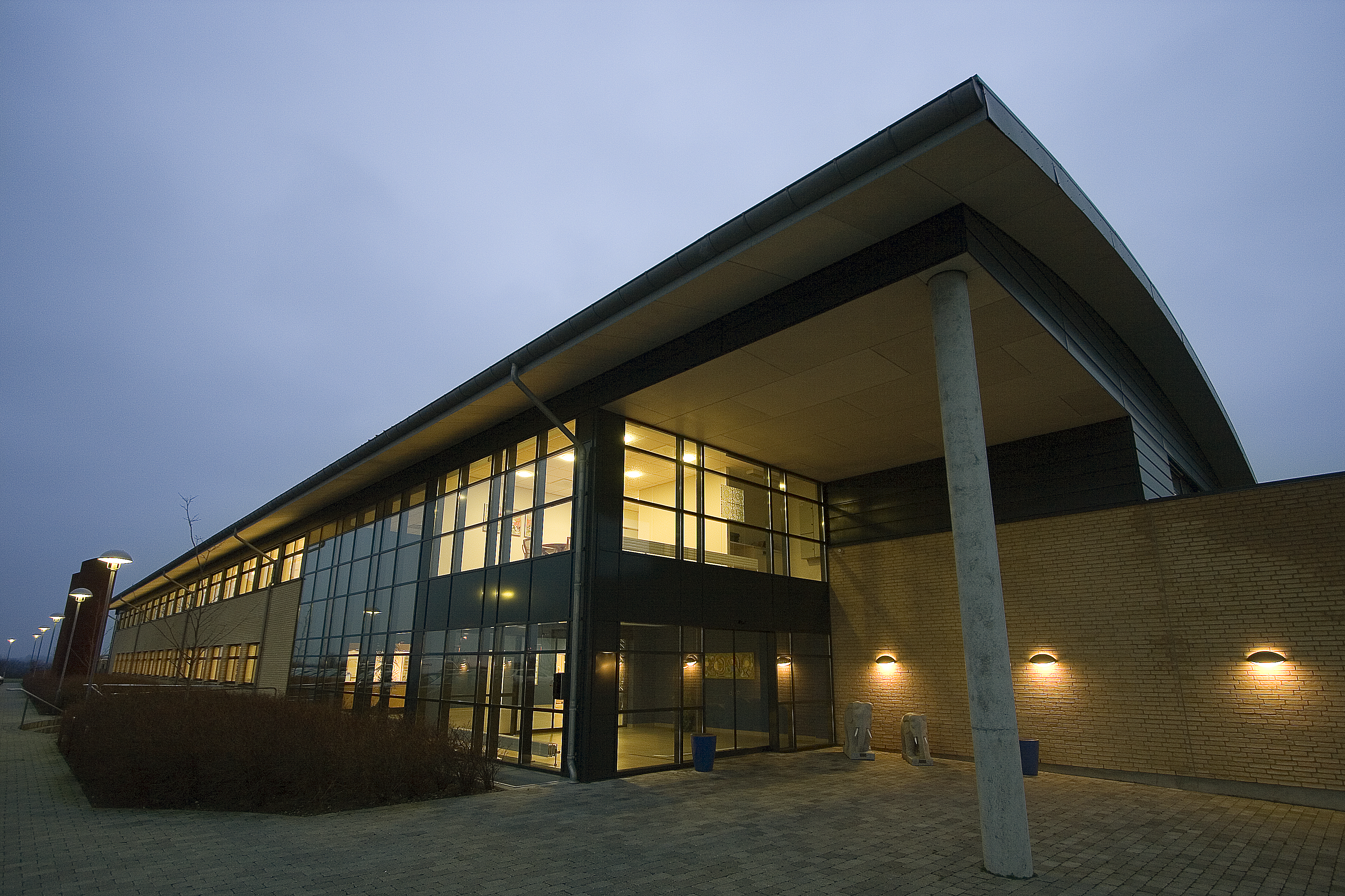 Modern building exterior with large glass windows and a sloped roof, illuminated under a twilight sky.