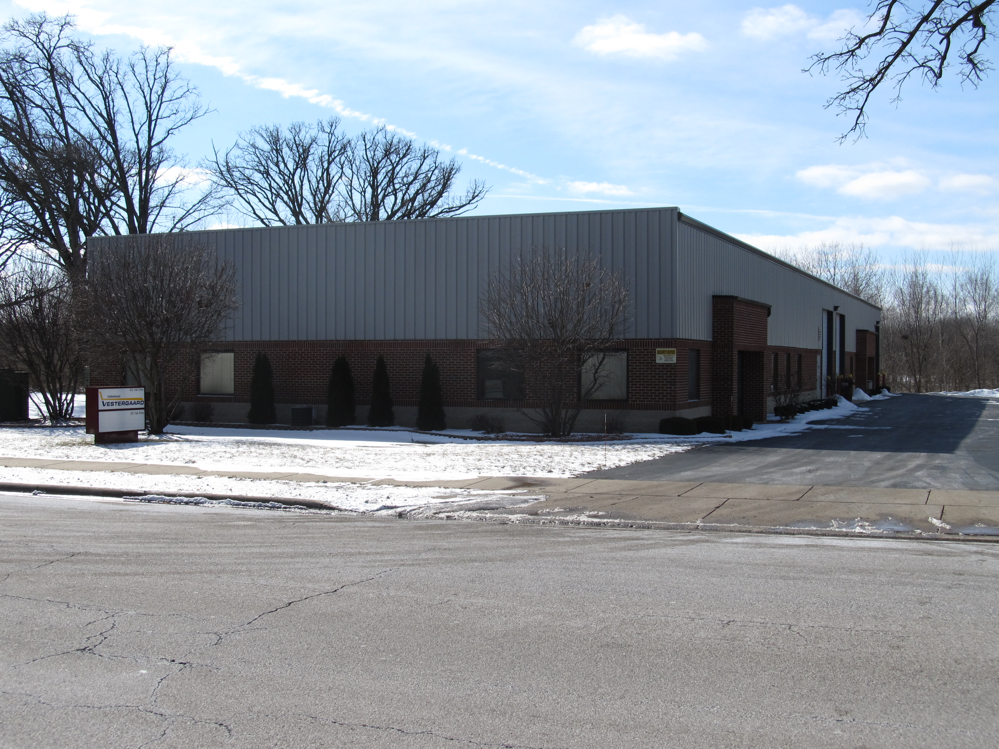 Single-story commercial building with a metal roof, surrounded by snow and trees, located on a paved road.