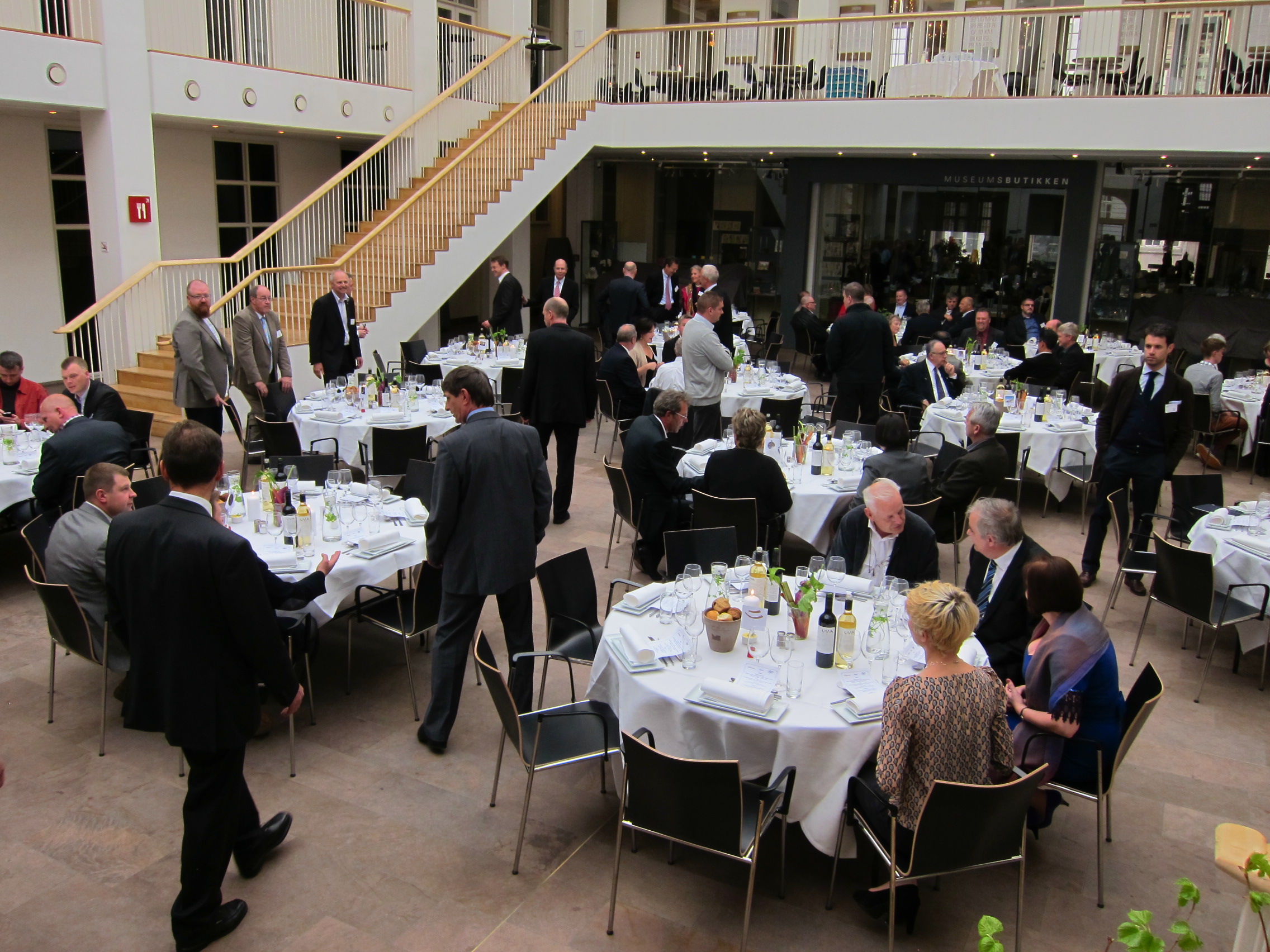 A formal gathering with numerous tables set for dining, attendees in suits and dresses engaged in conversation.