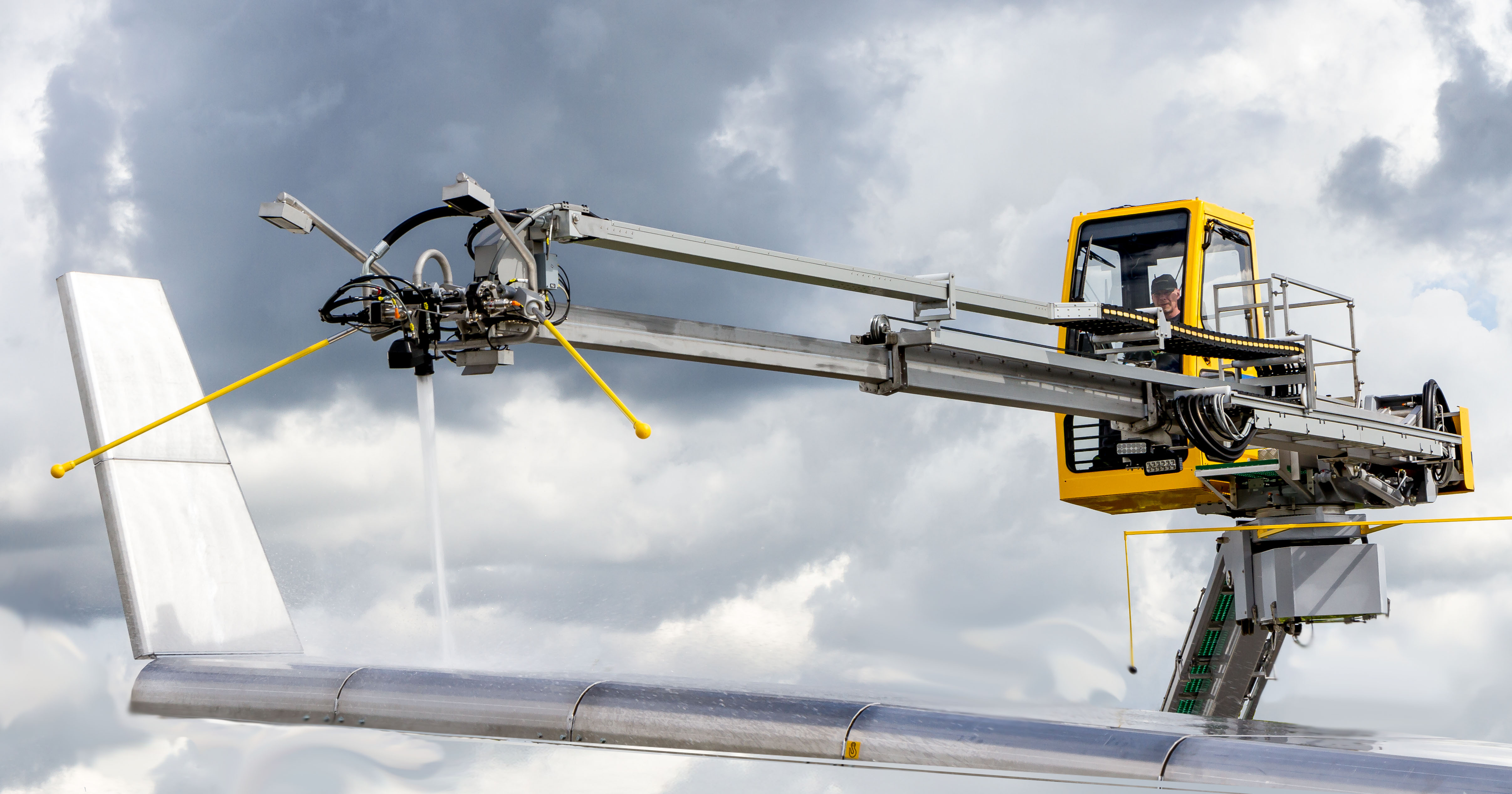 A yellow maintenance platform with a spray nozzle is positioned near an aircraft wing against a cloudy sky.