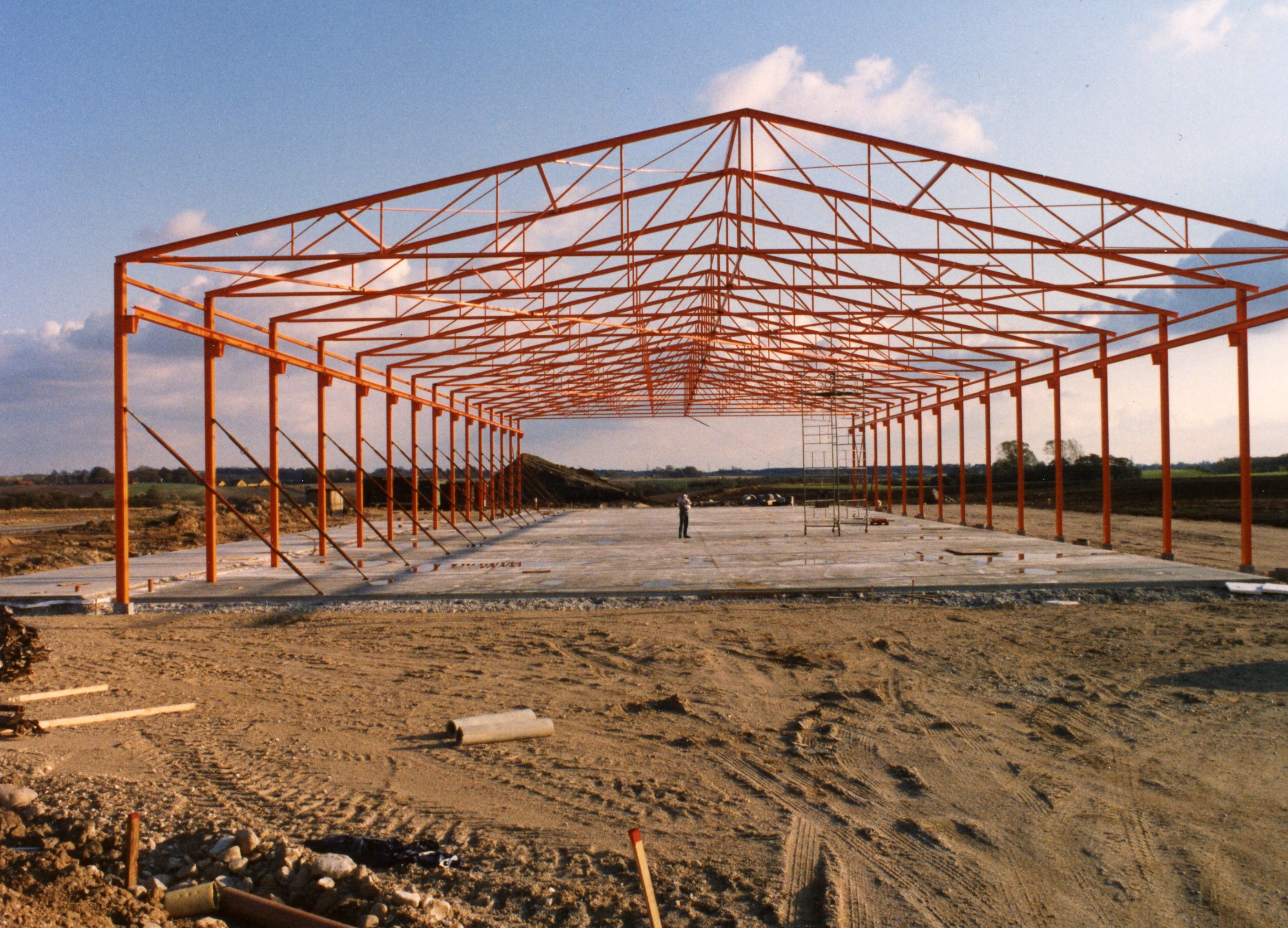Steel framework of a large building under construction, with a concrete foundation and a clear sky in the background.