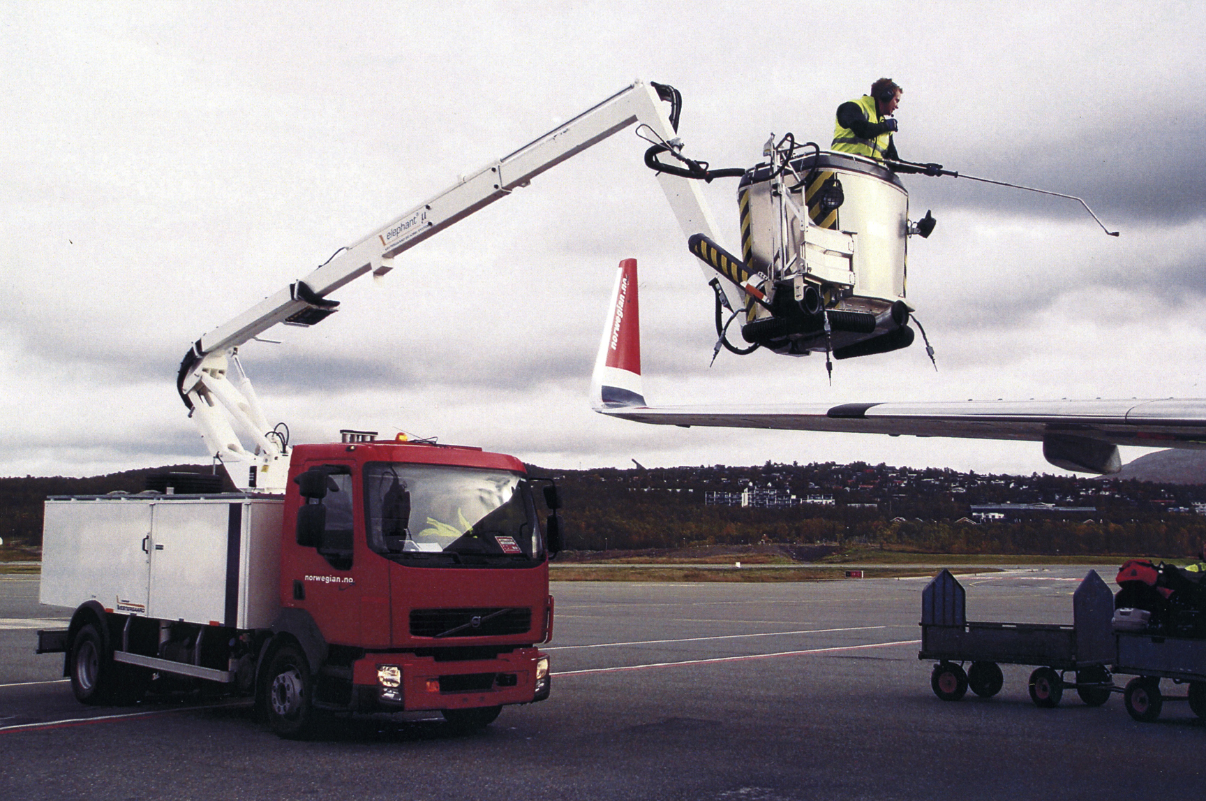 A worker in a lift performs maintenance on an airplane wing, with a red service truck nearby on the tarmac.