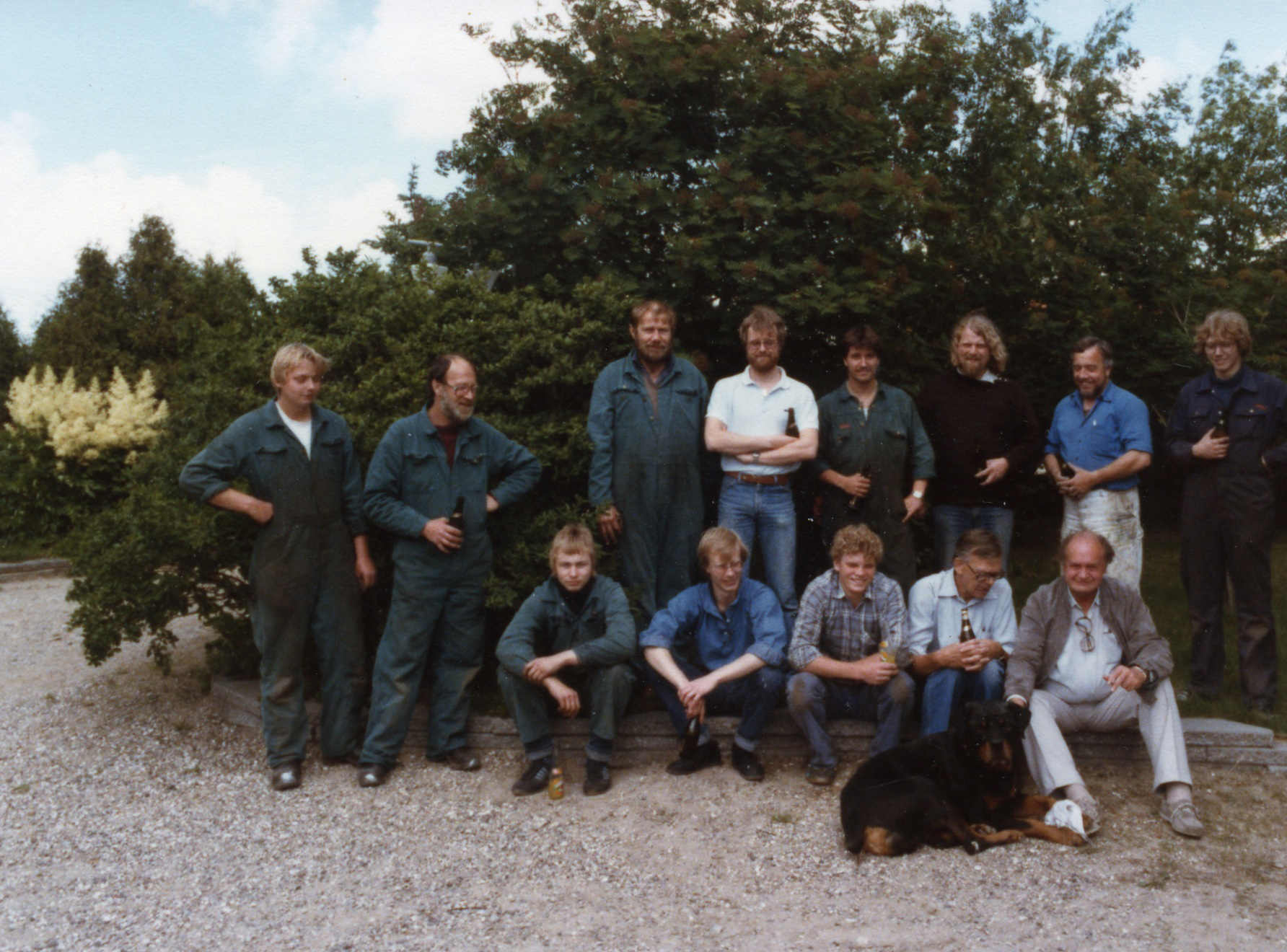 A group of 13 people in work attire, standing and sitting outdoors, with a dog in front, surrounded by trees.