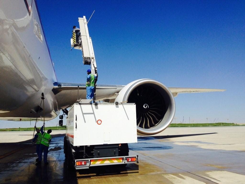 Two workers using a lift to service an aircraft engine on the tarmac under a clear blue sky.