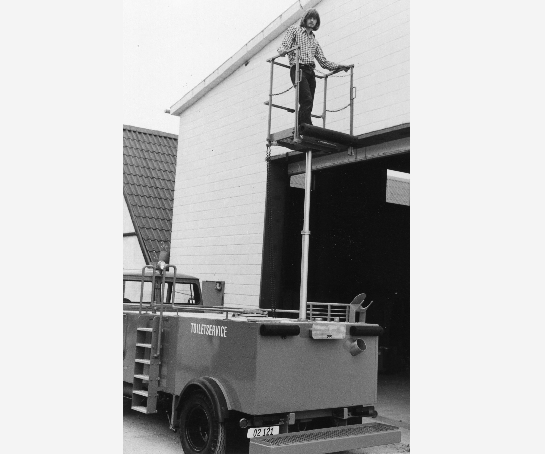 A worker stands on a lift platform, using a tool to reach the top of a building. A red utility vehicle is parked below.