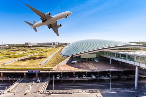 A commercial airplane flying above a modern airport terminal with a curved roof and clear blue sky.