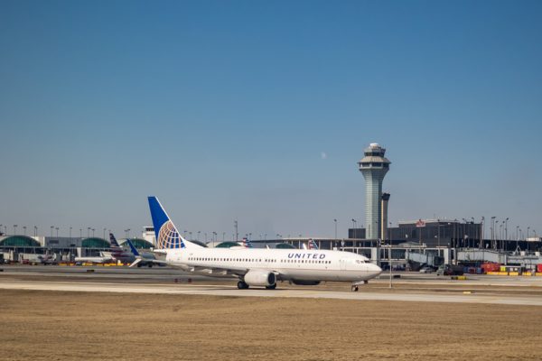 United Airlines aircraft taxiing on runway with airport control tower in background under clear blue sky.
