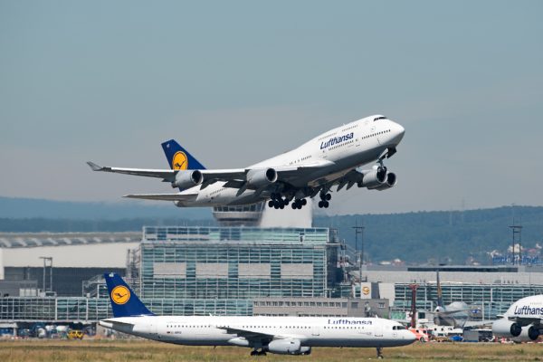 Lufthansa Boeing 747 taking off, with another Lufthansa aircraft on the runway and an airport terminal in the background.