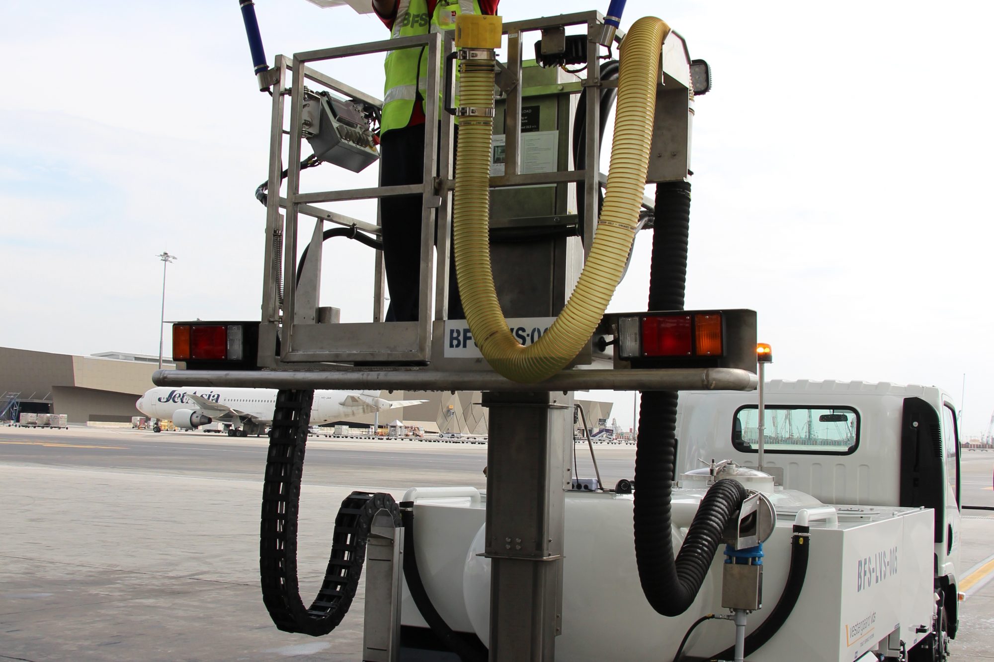A worker in safety gear operates a fuel truck with hoses connected to an aircraft for refueling on the tarmac.