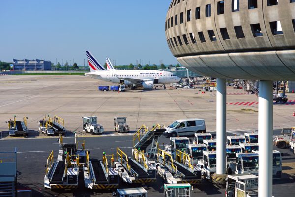 Airplane on runway with baggage carts and terminal building in foreground, clear blue sky in background.