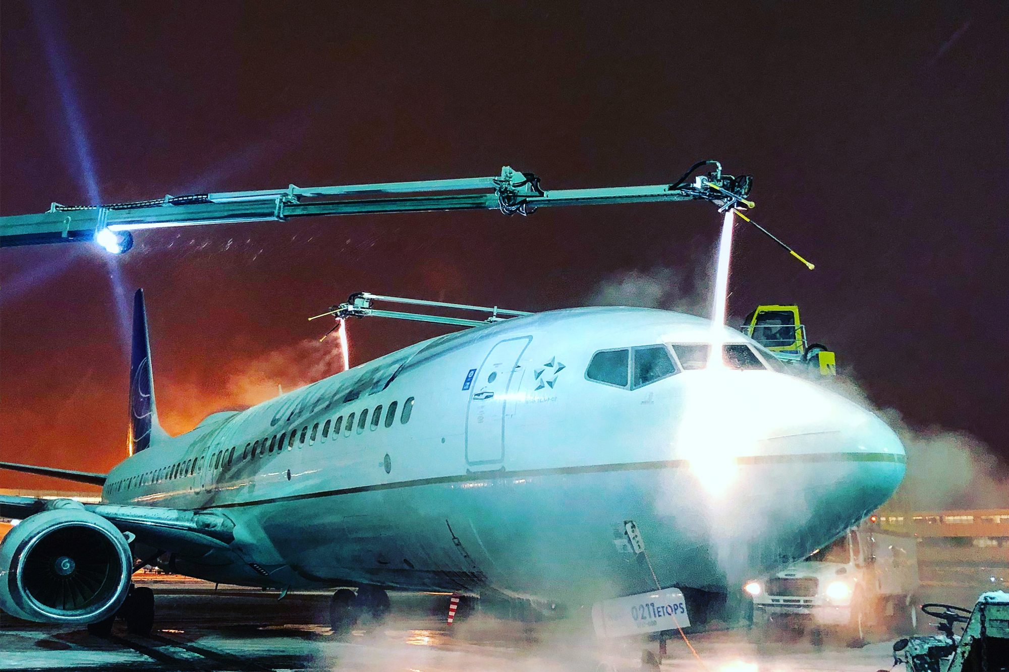 A commercial airplane being washed at night with water spraying from equipment, illuminated by blue lights.