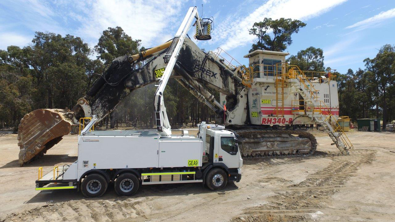 Large mining excavator with a white service truck underneath, both on a construction site surrounded by trees.