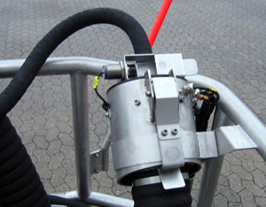 Mechanical component with a silver cylindrical body, attached hoses, and a red handle, likely part of a larger system.