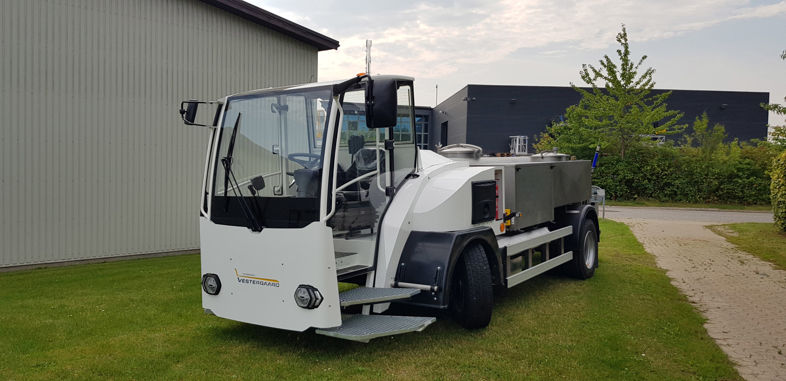 White industrial vehicle with a large tank, parked on grass near buildings.