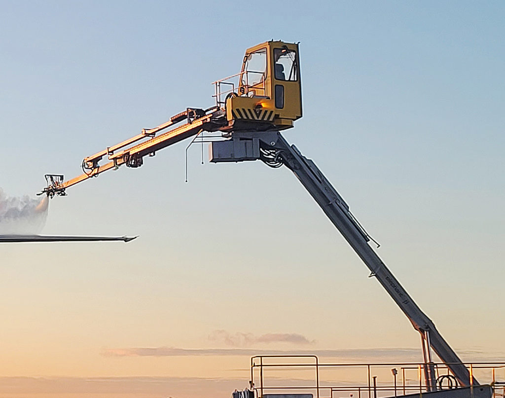 A yellow aerial lift with a long arm extending upwards against a sunset sky, used for maintenance or construction work.
