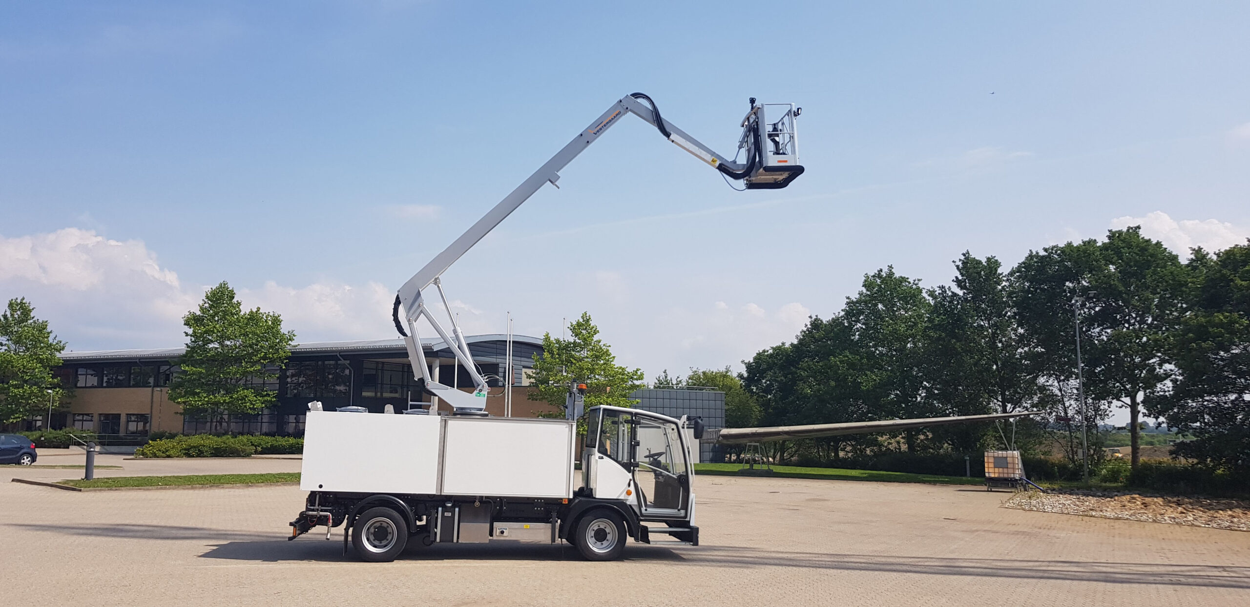 A truck with a raised aerial lift, positioned in a parking lot, surrounded by trees and buildings.