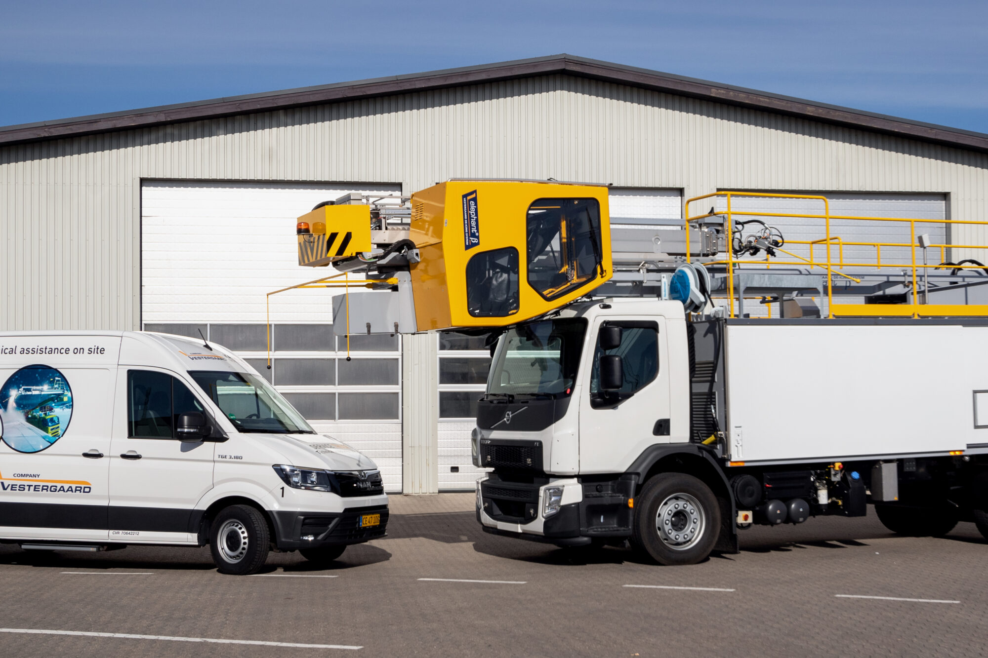 A white service van and a yellow and white truck with an elevated platform parked in front of a large building.