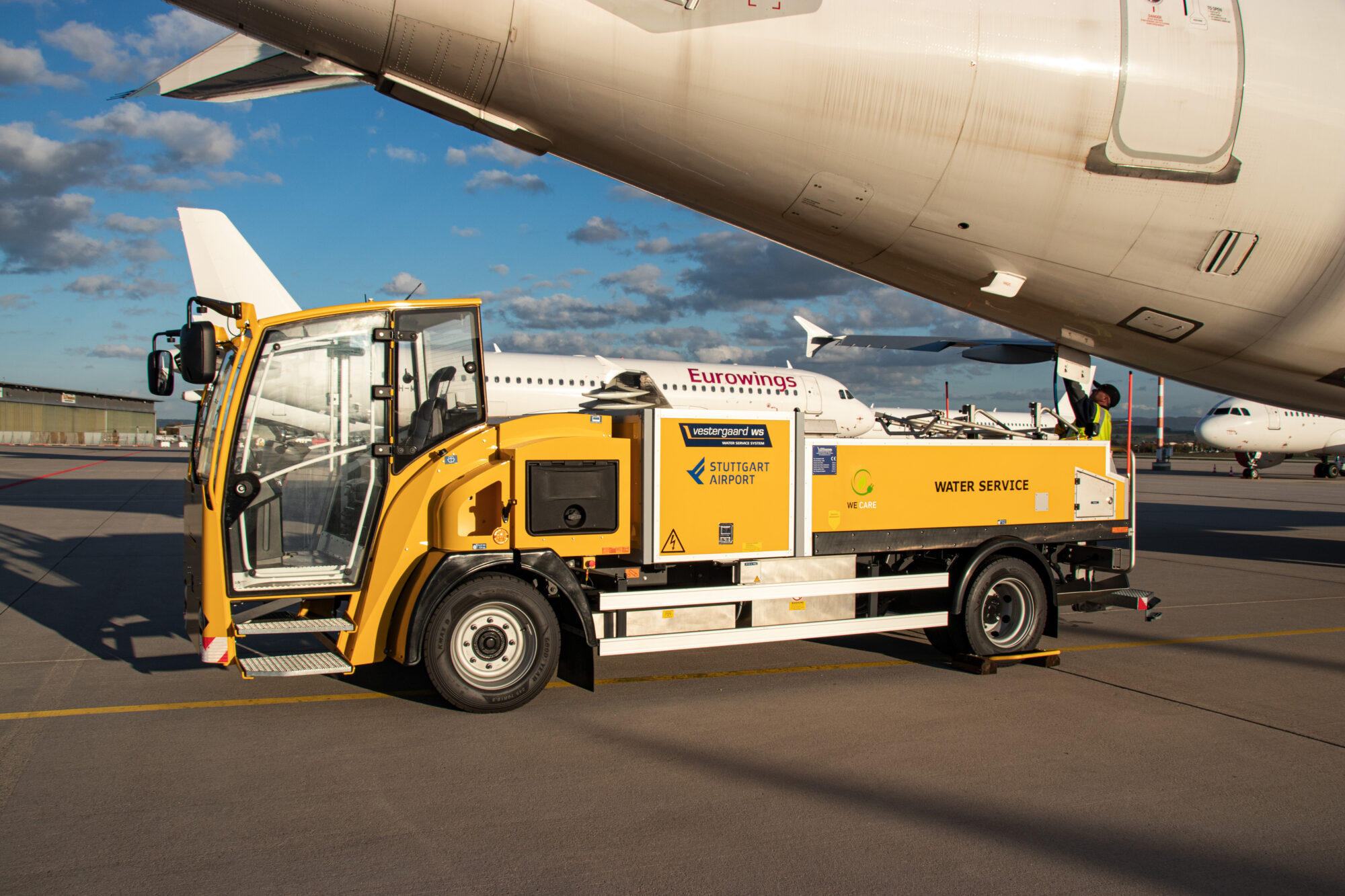Yellow aircraft servicing vehicle positioned under a plane, with clear skies and airport surroundings.