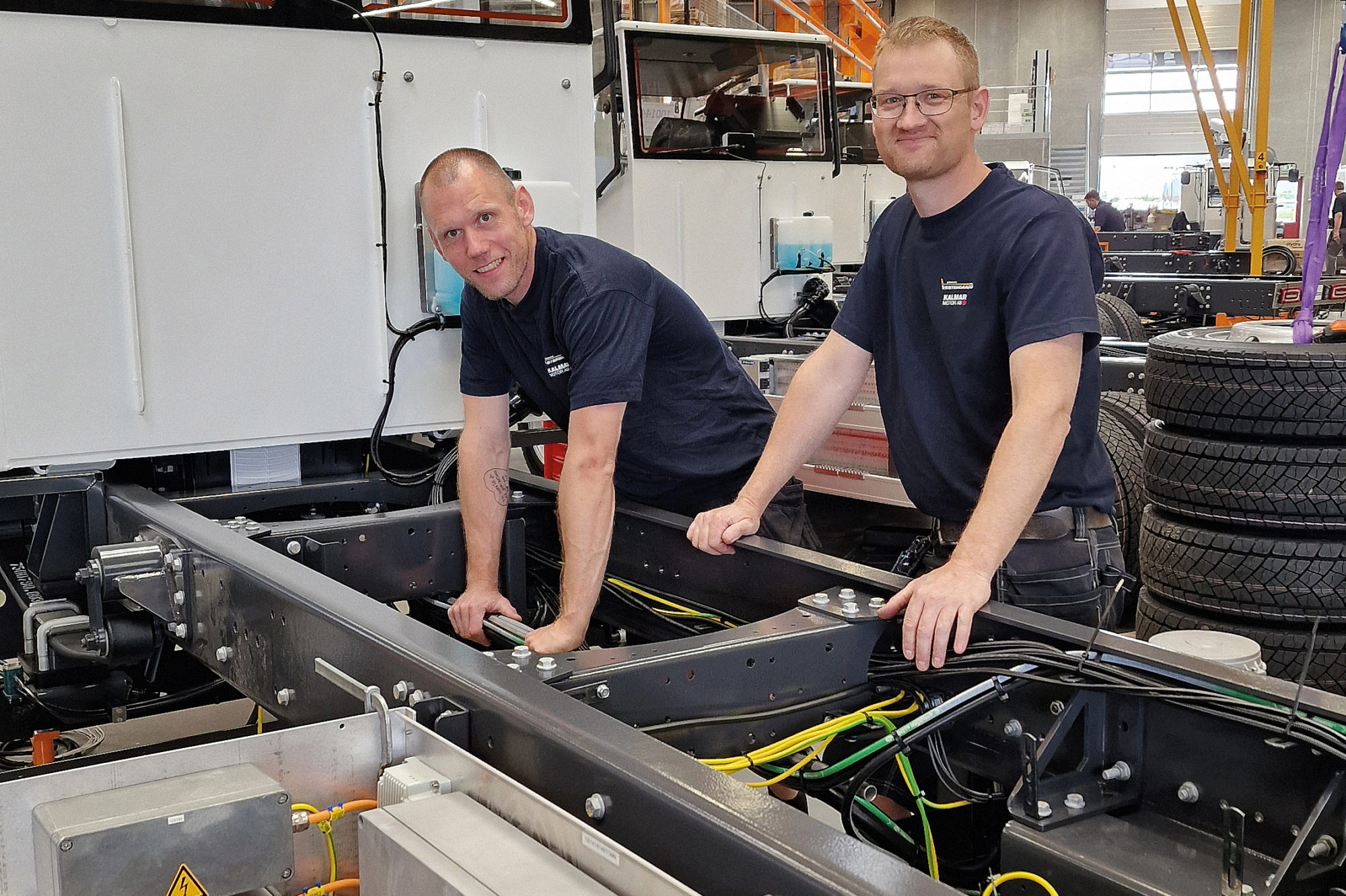 Two technicians working on a vehicle chassis, inspecting wiring and components in a workshop environment.