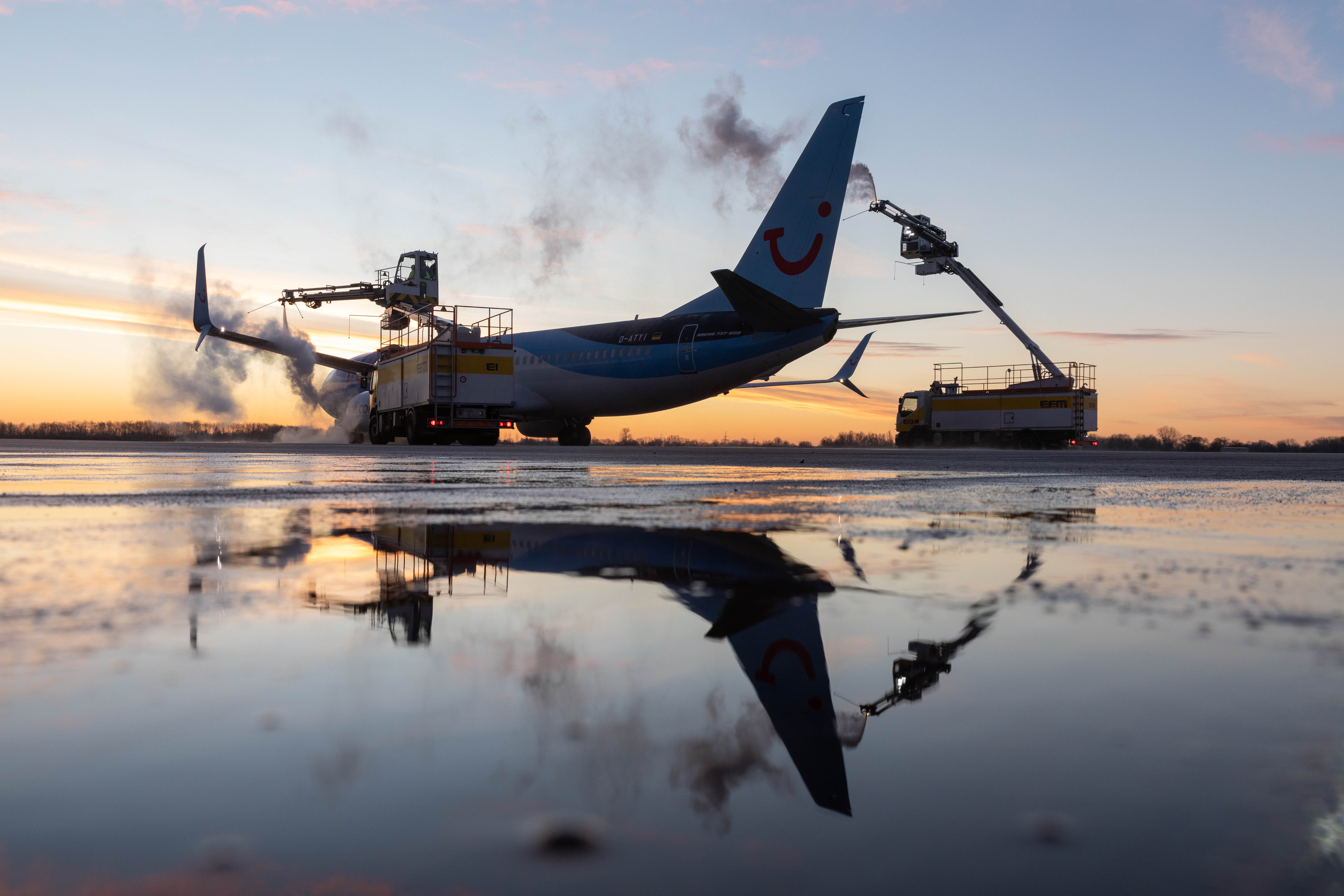 TUI aircraft being de-iced on the runway at sunset, with reflections in the water.